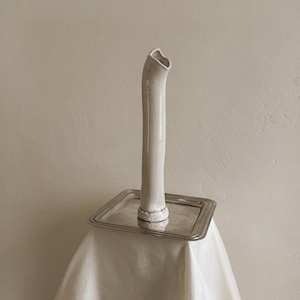 May include: A tall, slender, off-white ceramic vase with an irregular, organic cylindrical shape and a slightly flared, uneven rim. The vase is positioned upright in the center of a square, polished silver-toned metal tray with a thin, raised edge.