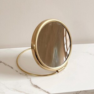 May include: A round, gold-toned compact mirror with a hinged design. The mirror is open, revealing the reflective surface. The frame is a bright gold color, and the base is a matching gold ring. The mirror is set against a neutral background.