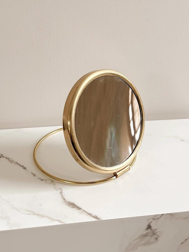 May include: A round, gold-toned compact mirror with a hinged design. The mirror is open, revealing the reflective surface. The frame is a bright gold color, and the base is a matching gold ring. The mirror is set against a neutral background.