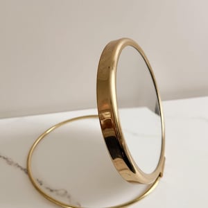 May include: A gold-toned, round compact mirror. The mirror is open, revealing the reflective surface. The frame and base are a matching gold color. The mirror is designed to stand upright on a circular base.