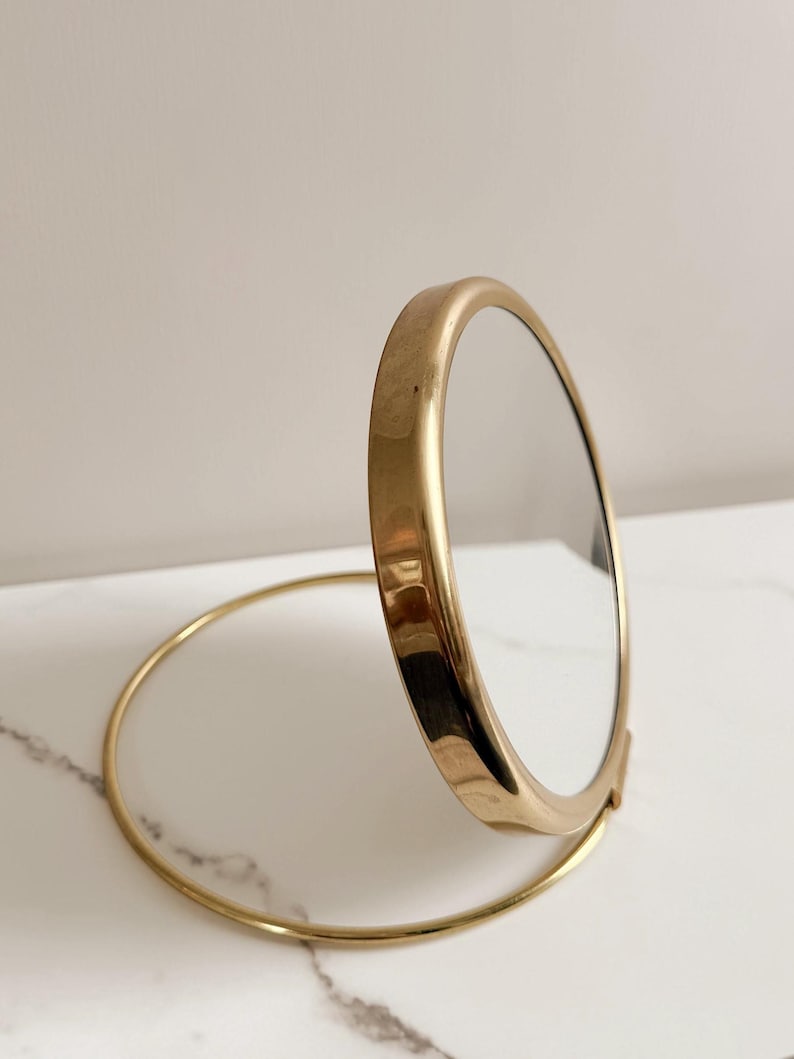 May include: A gold-toned, round compact mirror. The mirror is open, revealing the reflective surface. The frame and base are a matching gold color. The mirror is designed to stand upright on a circular base.