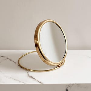May include: A round, gold-colored vanity mirror with a stand. The mirror is open, revealing the reflective surface. The stand is a thin, curved piece of metal. The mirror is on a white marble surface.