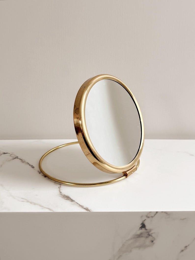 May include: A round, gold-colored vanity mirror with a stand. The mirror is open, revealing the reflective surface. The stand is a thin, curved piece of metal. The mirror is on a white marble surface.