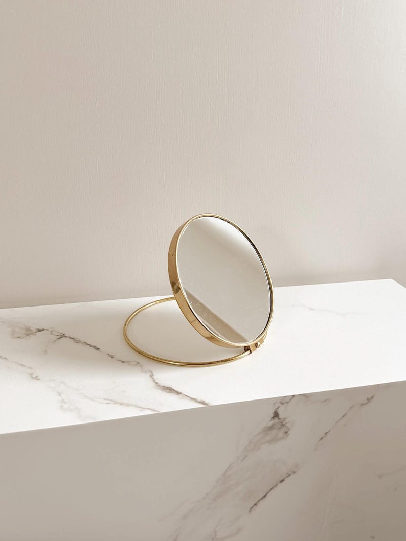 May include: A gold-colored, round, folding vanity mirror. The mirror is propped open on a white marble surface. The mirror's frame and base are a matching gold color.