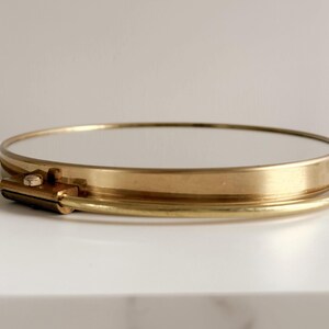 May include: A round, gold-toned metal tray with a mirrored surface. The tray has a simple, elegant design with a thin rim and a small clasp detail. The tray is sitting on a white surface.