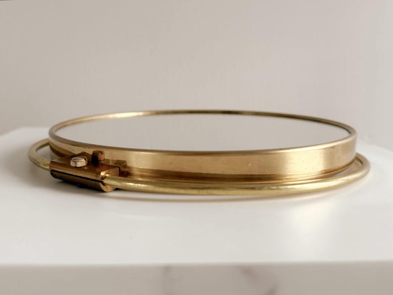 May include: A round, gold-toned metal tray with a mirrored surface. The tray has a simple, elegant design with a thin rim and a small clasp detail. The tray is sitting on a white surface.