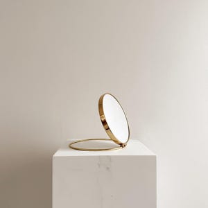 May include: A gold-colored, round compact mirror with a hinged design. The mirror is open, revealing the reflective surface. The mirror is set on a white, marble-like pedestal, creating a minimalist aesthetic.