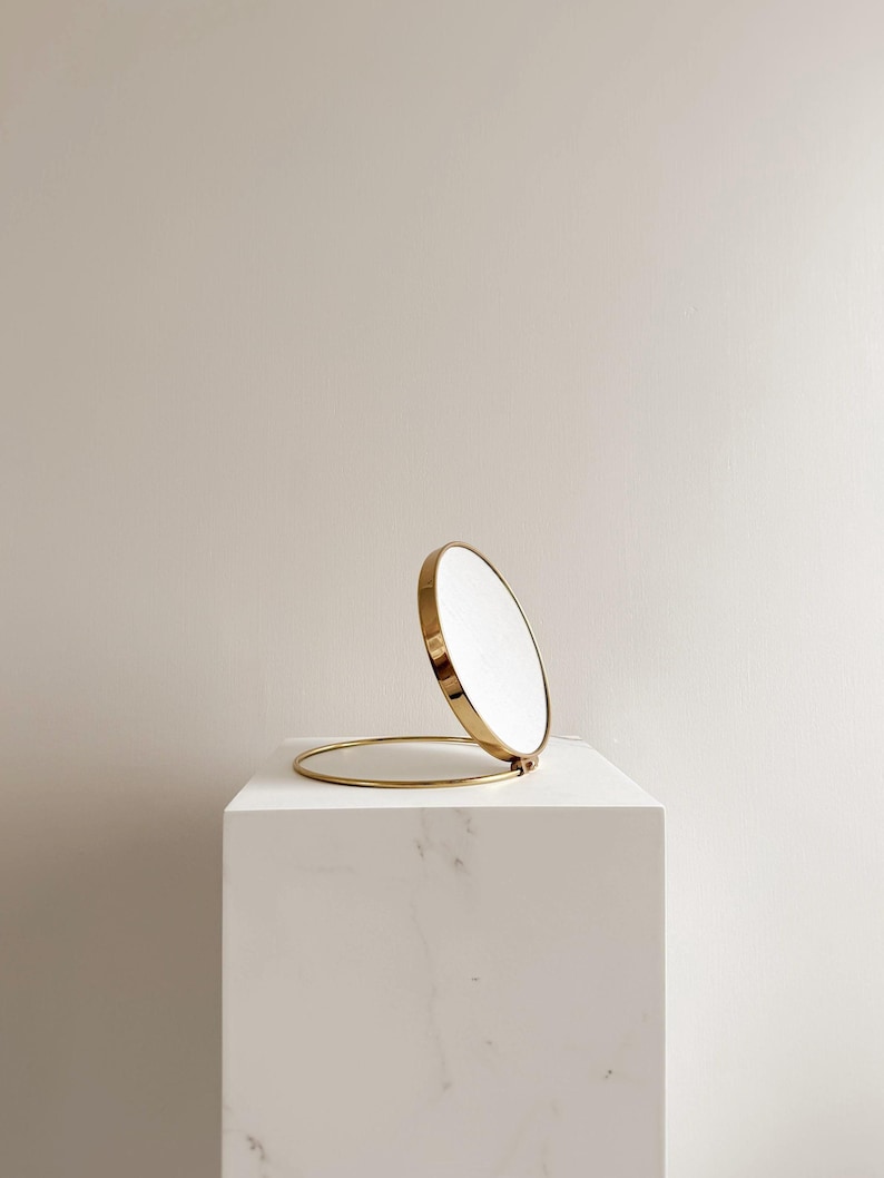 May include: A gold-colored, round compact mirror with a hinged design. The mirror is open, revealing the reflective surface. The mirror is set on a white, marble-like pedestal, creating a minimalist aesthetic.