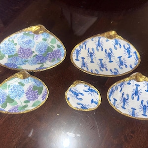 May include: A collection of five decorative seashell dishes. Two dishes feature a blue and purple floral pattern, while the other three display a blue lobster design on a white background. All shells have gold-painted edges.