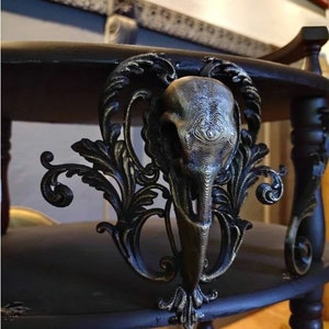 Può includere: Un tavolo in legno nero con una staffa metallica decorativa che presenta un design dettagliato di teschio di uccello. La staffa è fissata al bordo del tavolo e presenta elaborate volute.