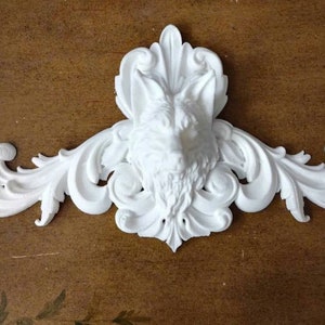 May include: A white decorative wall accent featuring a detailed fox head at the center, surrounded by ornate, swirling scrollwork. The design is symmetrical and made of a solid, smooth material, likely plaster or resin.