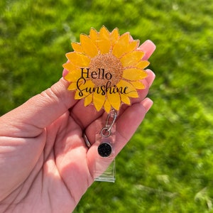 Pode incluir: Um carretel de crachá de plástico transparente com um botão preto de glitter e um girassol amarelo de glitter com o texto "Hello Sunshine".