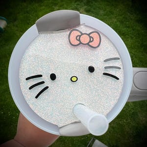 May include: White glitter tumbler lid with a Hello Kitty design. The lid has a grey silicone handle and a clear straw.
