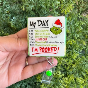 May include: A white and green badge reel with the text "My Day" and a schedule featuring the Grinch. The Grinch is wearing a Santa hat. The text "I'm Booked!" is in red. The reel has a clear plastic strap and a green button.