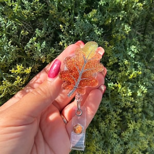 Puede incluir: Un portatarjetas con temática otoñal con un diseño de hoja de roble brillante en tonos naranja, dorado y plateado. La hoja está unida a un carrete de plástico transparente con un clip metálico. El portatarjetas se sostiene en una mano.
