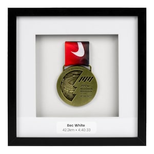 May include: A gold-colored medal with a Nike swoosh logo and the text "Melbourne Marathon Festival 02.10.2022" is displayed in a black frame with a white mat. The name "Bec White" and the text "42.2km • 4:40:33" are printed below the medal.