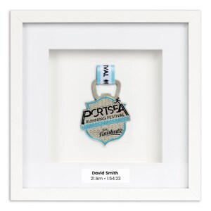 Könnte beinhalten: Gerahmte Medaille des Portsea Running Festivals mit hellblauem und silbernem Design. Die Medaille wird in einem weißen Rahmen mit dem Text "David Smith 21,1 km • 1:54:23" präsentiert.