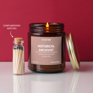 May include: A brown glass candle jar with a gold lid. The candle is lit and the label reads "VEYS GIFT SHOP HISTORICAL ARCHIVIST smells like old documents and past eras. 207g - 7.2 oz SCENTED SOY CANDLE". A small glass jar with a cork lid filled with matches is next to the candle.