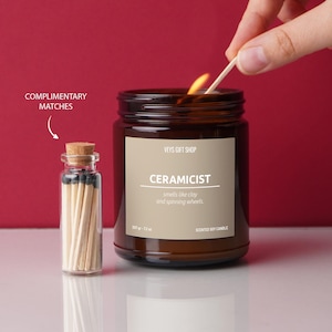 May include: A lit brown glass jar candle with the text "CERAMICIST" and a small glass bottle of matches. The candle is being lit with a match. The candle smells like clay and spinning wheels.