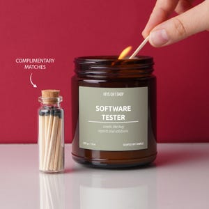 May include: A brown glass candle jar with a white label that reads "VEYS GIFT SHOP" and "SOFTWARE TESTER" with the tagline "smells like bug reports and solutions." The candle is lit and a matchstick is being used to light it. A small glass jar with matches is next to the candle.
