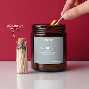 May include: A brown glass candle jar with a white label that reads "VEYS GIFT SHOP" and "ARCHITECT" in a bold font. The label also says "smells like drafting paper and visionary ideas." The candle is lit and there is a small glass jar of matches next to it.