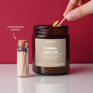 May include: A brown glass candle jar with a white label that reads "FUNERAL DIRECTOR" and "smells like solemn respect and final goodbyes." The candle is lit and there is a small jar of matches next to it.