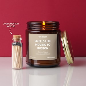 May include: A brown glass candle jar with a gold lid. The candle is lit and the jar has a label that reads "Smells Like Moving to Boston. The city of history and academic brilliance. Scented Soy Candle". A small glass jar with matches is included.