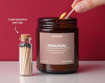 Może przedstawiać: Szklany słoik w kolorze brązowym z woskową świecą, na której widnieje etykieta z napisem "VEYS GIFT SHOP" i "PARALEGAL smells like legal documents and critical thinking." Świeca jest zapalona, a do jej zapalenia użyto zapałki. Obok świecy znajduje się mały szklany słoik z zapałkami.