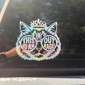 May include: An iridescent holographic decal featuring a stylized cat wearing a crown and round glasses. The text "THIS IS AN OUT RAGE!" is printed inside the lenses of the glasses. The sticker is applied to a dark glass surface, showing a reflective finish.