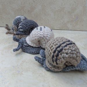 May include: Three handmade crochet snails in a row. The snails have gray, beige, and off-white shells with dark gray stripes and accents. The bodies are gray and beige. The snails are on a light-colored surface.