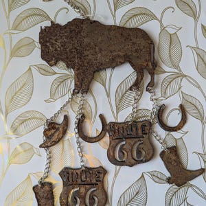 May include: A rustic metal Route 66 themed wall hanging. The design features a bison, cowboy boots, horseshoes, and Route 66 signs, all connected by chains. The metal has a weathered, brown finish, giving it a vintage look. The background has a gold and white leaf pattern.