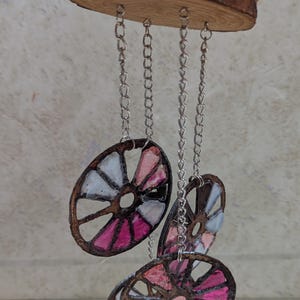 May include: A handcrafted wind chime featuring a natural wood slice base with multiple decorative, stained glass-style circular elements. The circles are in shades of pink, white, and gray, suspended by silver chains.