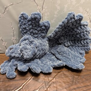 Peut inclure: Un dragon en peluche fait à la main, d'un bleu doux et sourd. Le dragon présente de grandes ailes détaillées, une petite tête et quatre pattes griffues. Le jouet est fait d'une matière texturée et veloutée, lui donnant un aspect câlin. Le dragon repose sur une surface en bois.