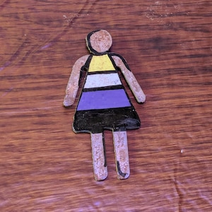 May include: A handmade wooden figure in the shape of a person, painted with the colors of the non-binary pride flag: yellow, white, purple, and black. The figure has a simple, stylized design with a head, torso, arms, and legs.