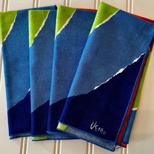 May include: Four folded cloth napkins with a modern design. The napkins feature a color block design with shades of blue, green, and white. The bottom of the napkins are a deep navy blue. The napkins are stacked on a white surface. The signature "Ueia" is visible.