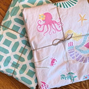 May include: Two sets of bedding, one with a teal geometric pattern and the other with a pink sea-themed design. The pink set features an octopus, mermaid, starfish, and other sea creatures. Both sets are tied with twine.