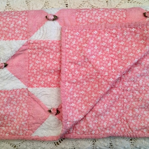 May include: A pink and white patchwork quilt with a floral pattern. The quilt is decorated with pink fabric flowers.