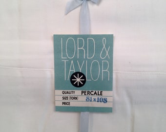 May include: A white sheet with a blue ribbon tied in a bow. A blue tag with the text "LORD & TAYLOR" and "PERCALE" is attached to the ribbon. The tag also includes the text "QUALITY", "SIZE (TORN)", "PRICE", and "81x108".