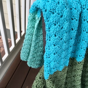 May include: A crocheted blanket with a gradient of green, teal, and blue colors. The blanket is folded and draped over a chair.