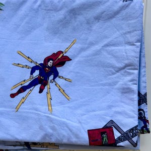 May include: Light blue Superman-themed bed sheet featuring a large graphic of Superman in flight with yellow lightning bolts. The sheet also has smaller illustrations of Superman and other graphics.