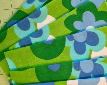 May include: A stack of fabric with a retro floral pattern. The design features large, stylized flowers in shades of green, blue, and white. The fabric has a textured appearance, and the flowers are arranged in a repeating pattern.