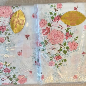 May include: Two packages of white percale sheets and pillowcases with a floral pattern. The sheets and pillowcases are decorated with pink roses and green leaves. The packages are sealed in clear plastic and have gold labels.