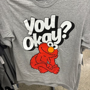 May include: Gray t-shirt with a red Sesame Street character, Elmo, sitting on the bottom of the shirt. The text "you okay?" is printed in white on the shirt.