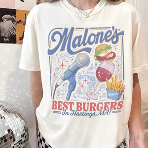 May include: A cream-colored short-sleeve t-shirt featuring a graphic print with the text Malone's BEST BURGERS In Hastings, MA! The design includes a blue microphone, a burger, a glass of red wine, and a container of french fries in blue and red.
