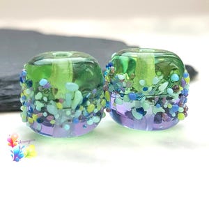 May include: Two cylindrical glass beads featuring a gradient of translucent green at the top and purple at the bottom. The exterior is decorated with a textured band of small, multi-colored glass frit in shades of blue, green, and purple.