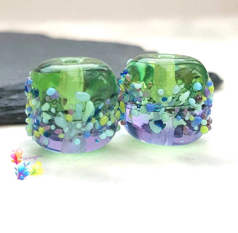 May include: Two cylindrical glass beads featuring a gradient of translucent green at the top and purple at the bottom. The exterior is decorated with a textured band of small, multi-colored glass frit in shades of blue, green, and purple.