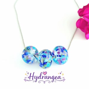 May include: A silver necklace with three glass beads in blue, purple, and green. The beads are spherical and have a floral pattern. The word "Hydrangea" is written in a decorative font below the necklace.