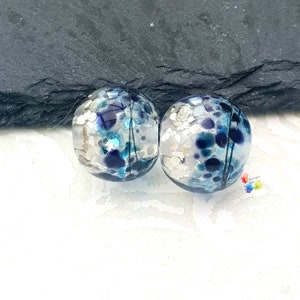 Könnte beinhalten: Zwei runde Glasperlen mit einem Mosaik aus Silber, Blau und Türkis. Die Perlen haben ein gesprenkeltes Aussehen mit dunkelblauen Akzenten. Die Perlen werden auf einer dunkelgrauen Oberfläche vor einem weißen Hintergrund präsentiert.