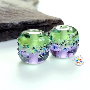May include: Two cylindrical glass beads featuring a horizontal color split of lime green on top and lavender purple on the bottom. A textured band of small, multi-colored glass frit particles encircles the center. Text reads Glittering Prize Colouring Your World.