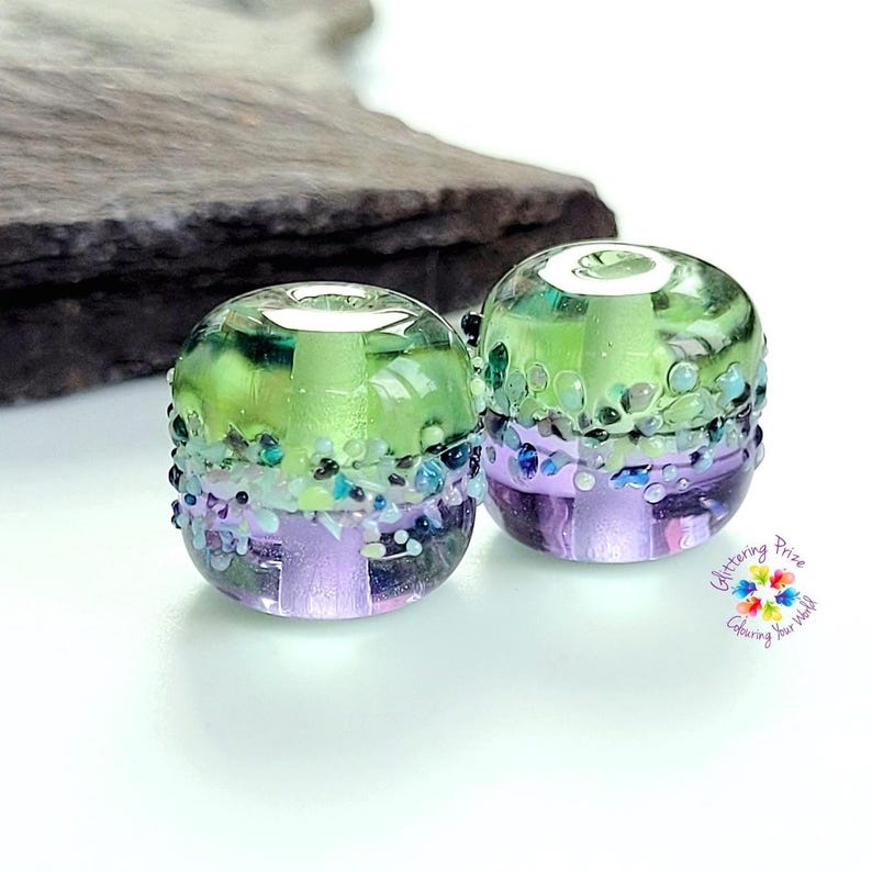 May include: Two cylindrical glass beads featuring a horizontal color split of lime green on top and lavender purple on the bottom. A textured band of small, multi-colored glass frit particles encircles the center. Text reads Glittering Prize Colouring Your World.